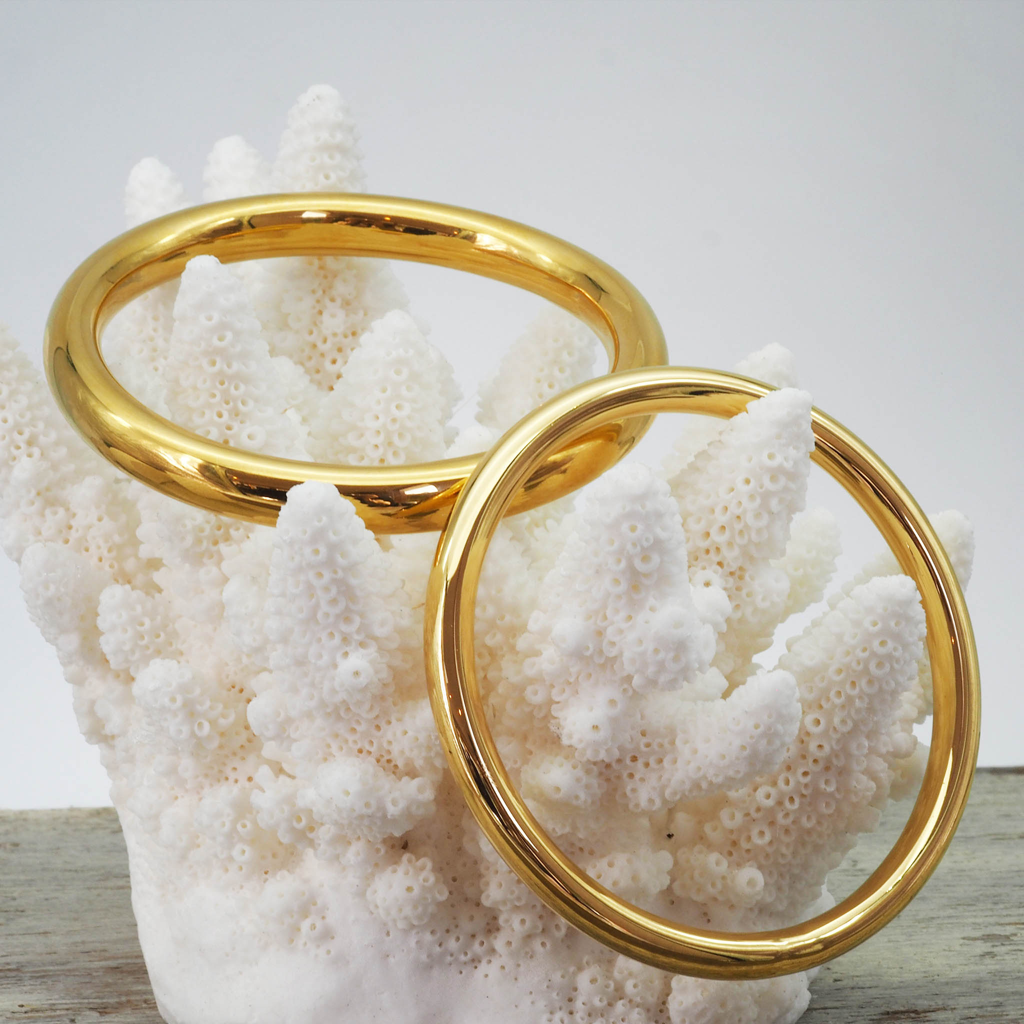 two gold stainless steel bangle bracelets in polished finish on a coral