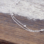 A delicate silver double chain anklet with small polished bead accents, displayed on a rustic wooden surface.