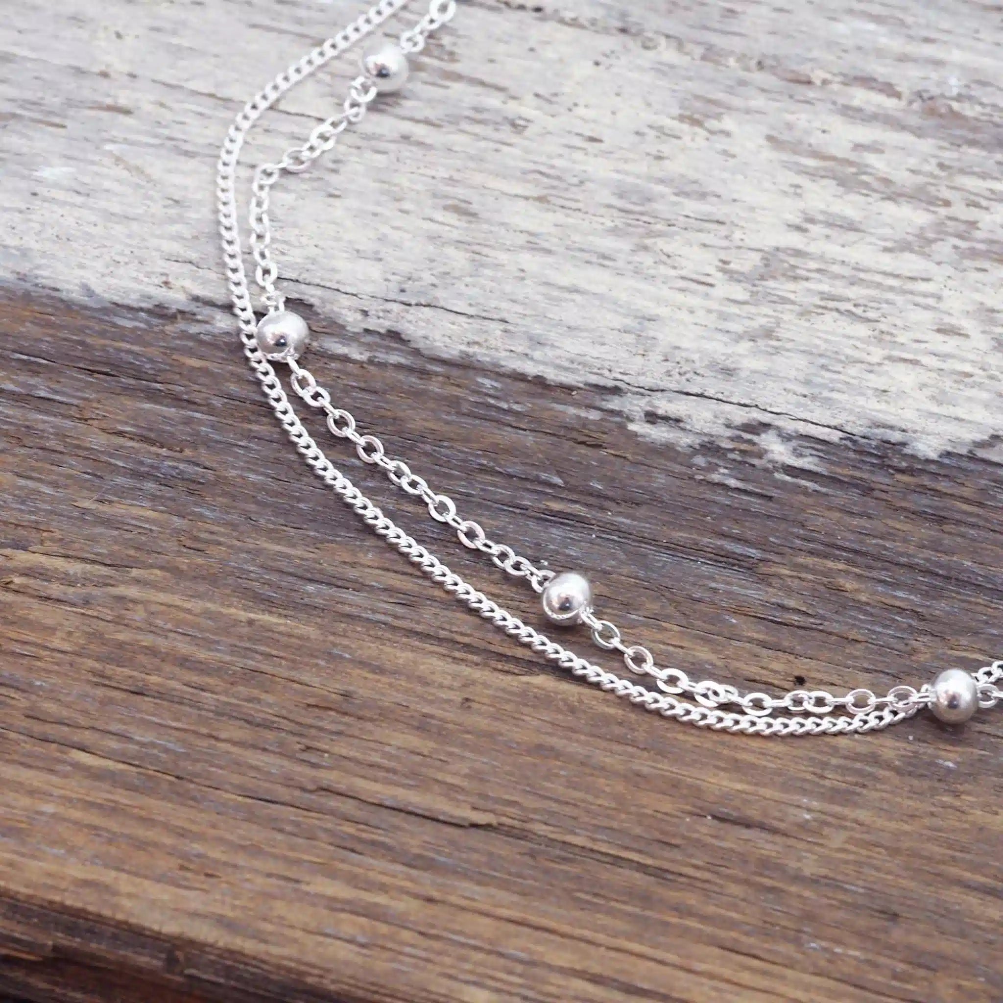 A delicate silver double chain anklet with small polished bead accents, displayed on a rustic wooden surface.