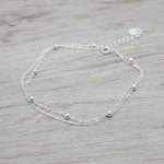 A delicate double-layered silver anklet with small bead accents, displayed on a light gray wooden surface.