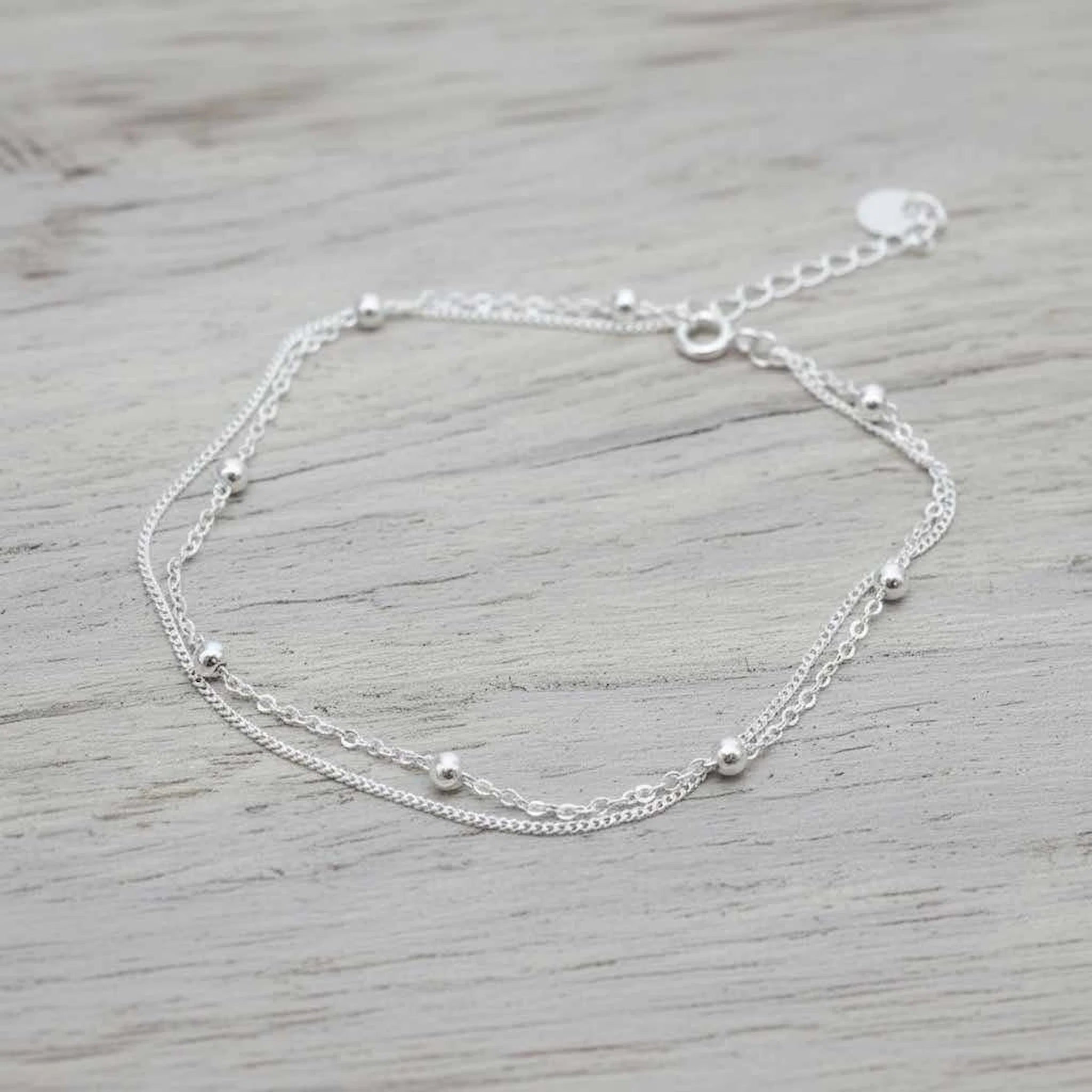 A delicate double-layered silver anklet with small bead accents, displayed on a light gray wooden surface.