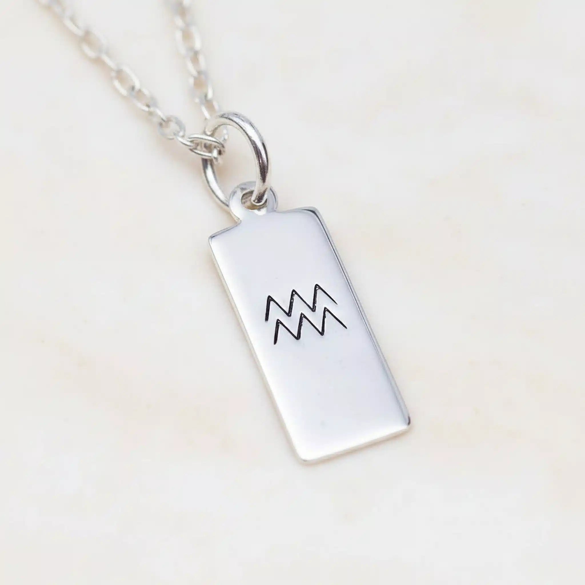Silver necklace with a rectangular pendant featuring an Aquarius symbol on a light beige background