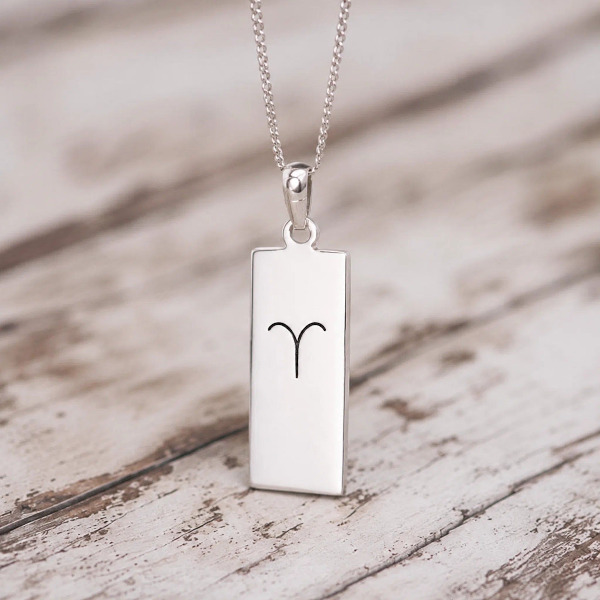 A silver rectangular pendant necklace engraved with the Aries zodiac symbol, resting on a rustic, light-colored wooden surface.