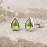A pair of silver August Birthstone Natural Peridot Earrings - teardrop stud earrings with smooth olive green gemstone centers, displayed on a textured beige stone surface with small dried flowers in the background.