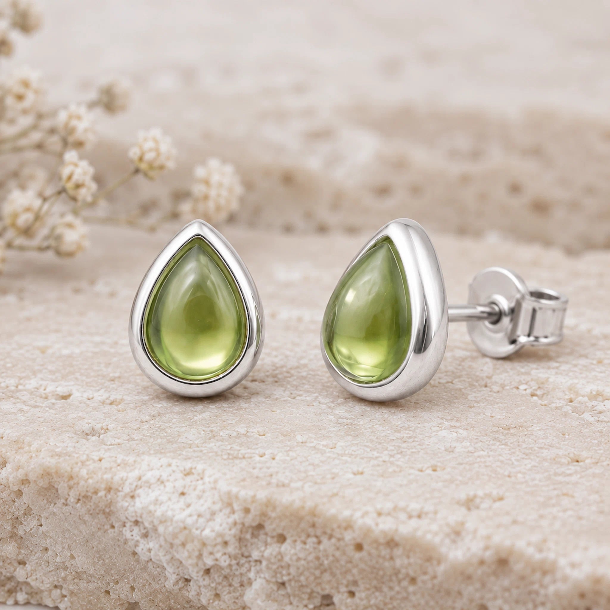 A pair of silver August Birthstone Natural Peridot Earrings - teardrop stud earrings with smooth olive green gemstone centers, displayed on a textured beige stone surface with small dried flowers in the background.