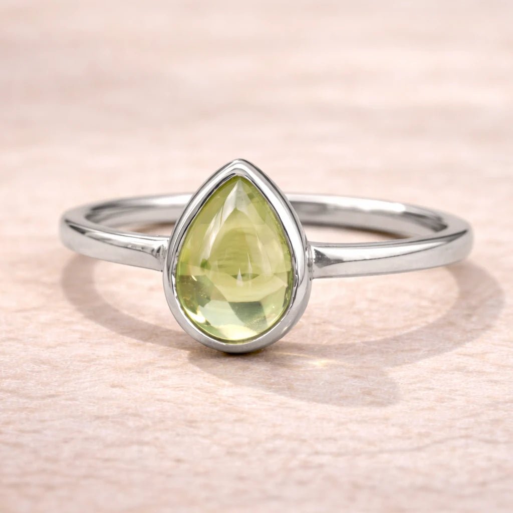 Sterling Silver August birthstone ring with a pear-shaped light green natural peridot gemstone cabochon in a polished low-profile bezel setting on a beige stone surface