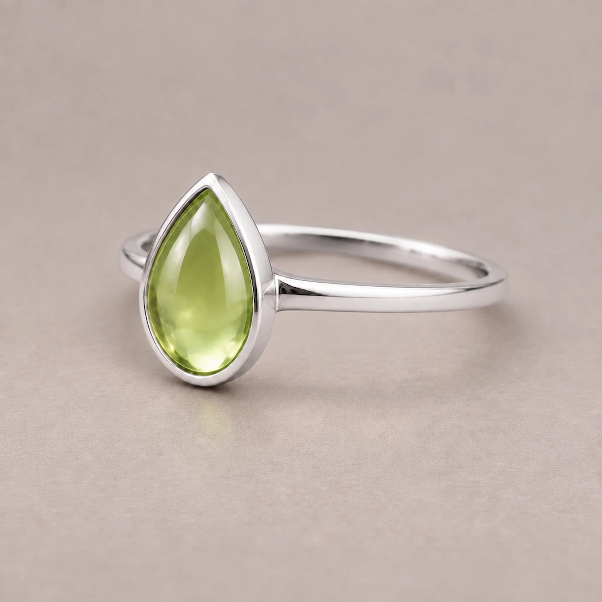 Sterling Silver August birthstone ring with a pear-shaped light green natural peridot gemstone cabochon in a polished low-profile bezel setting on a beige surface