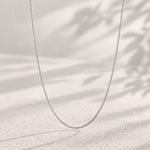 A delicate silver chain necklace draped in a U-shape against a warm, textured beige background with soft leaf shadows.