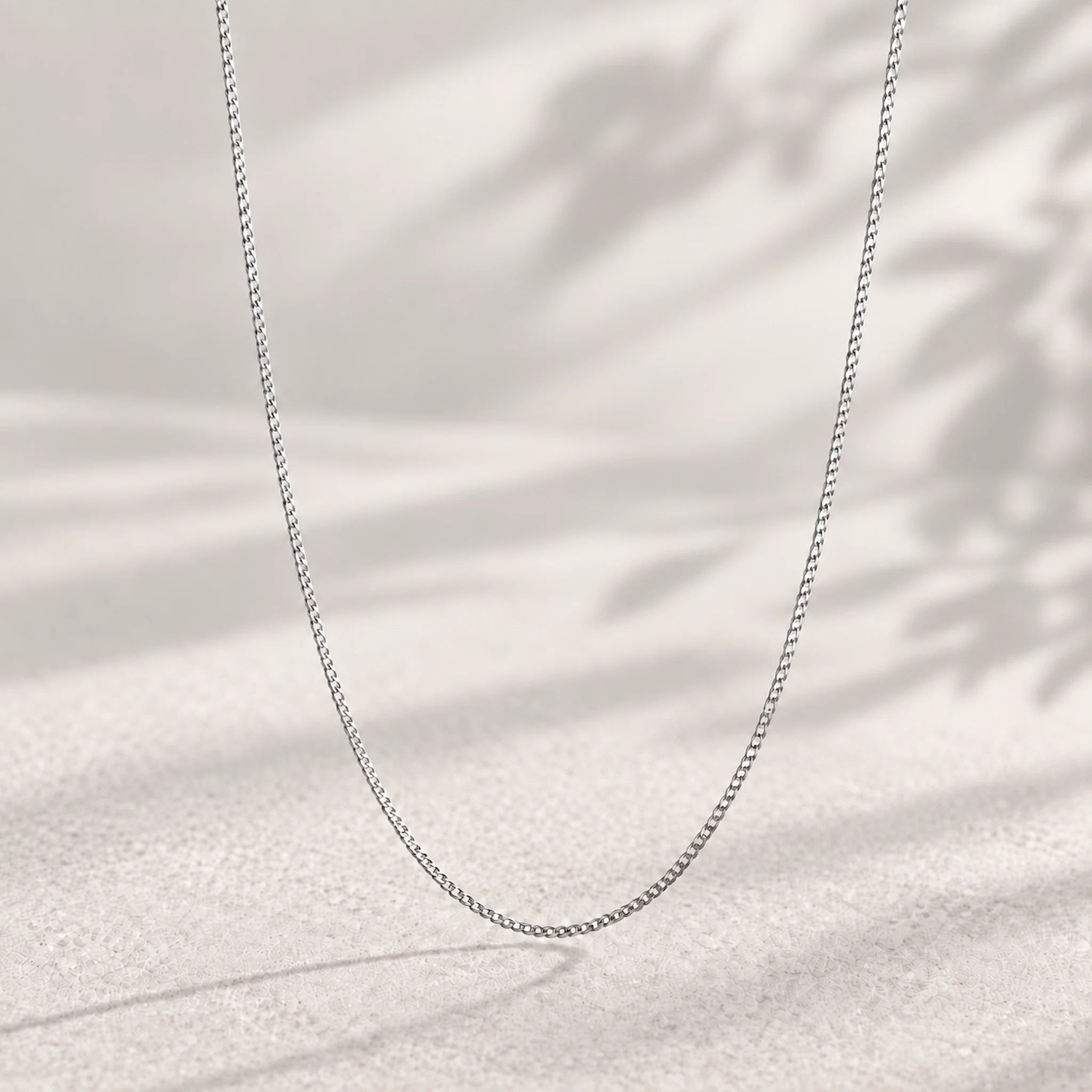 A delicate silver chain necklace draped in a U-shape against a warm, textured beige background with soft leaf shadows.