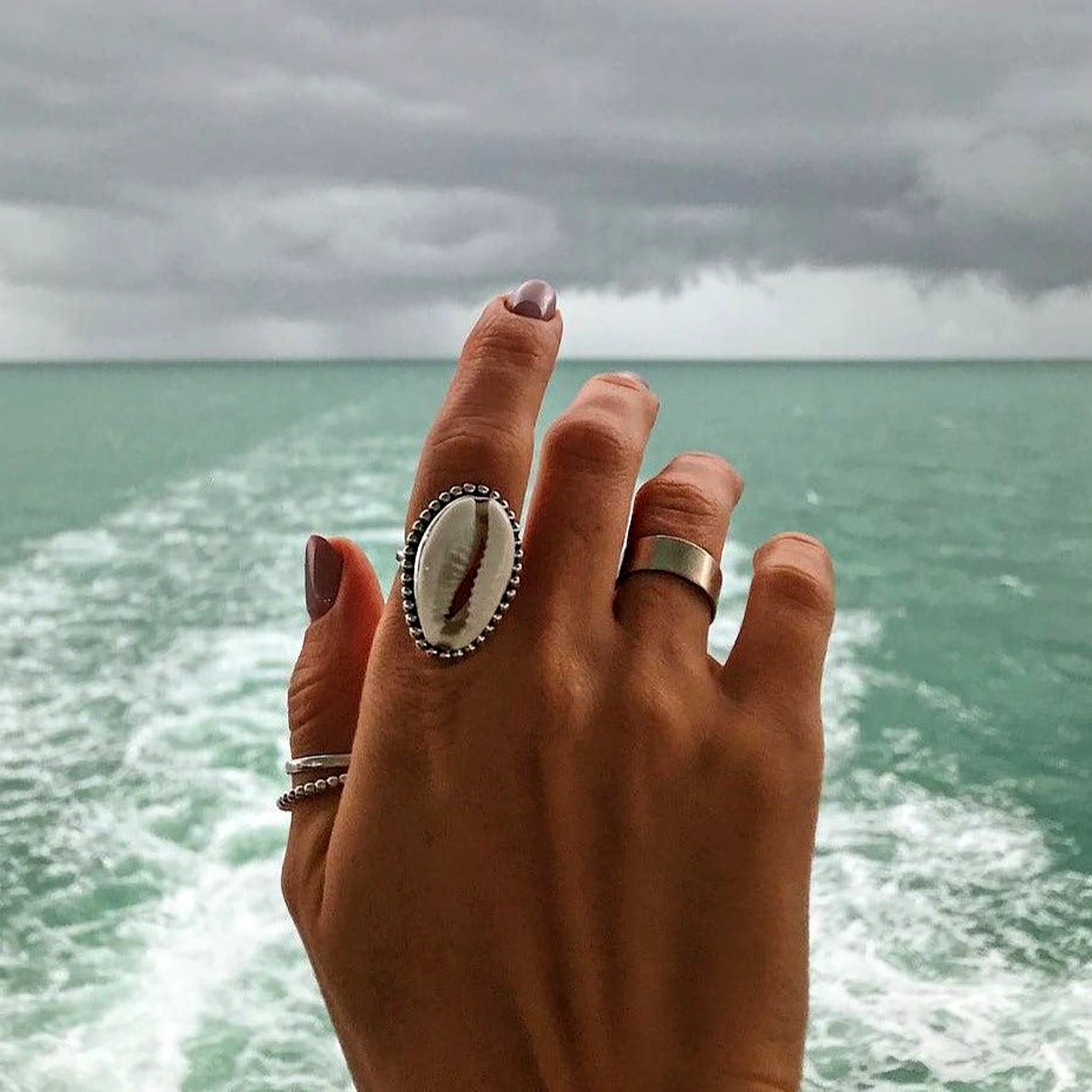 Hand wearing a ring with a shell design against a backdrop of ocean waves and cloudy sky.