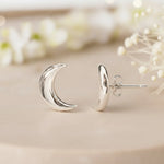 Silver crescent moon earrings on a neutral background with blurred flowers.
