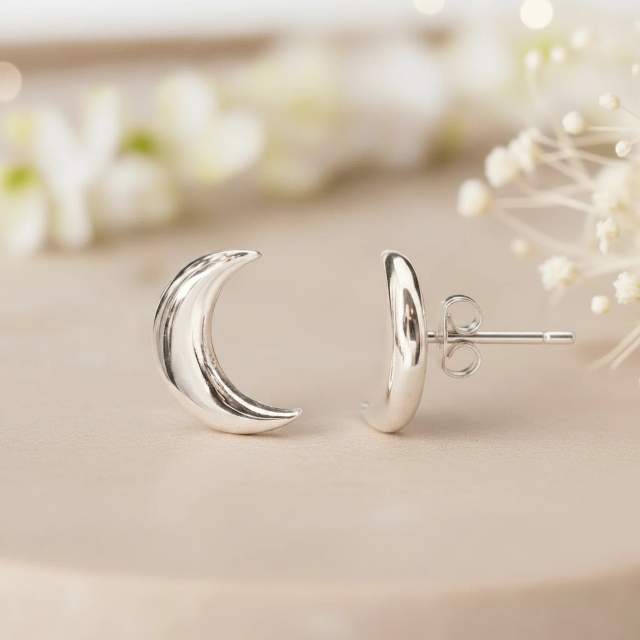 Silver crescent moon earrings on a neutral background with blurred flowers.