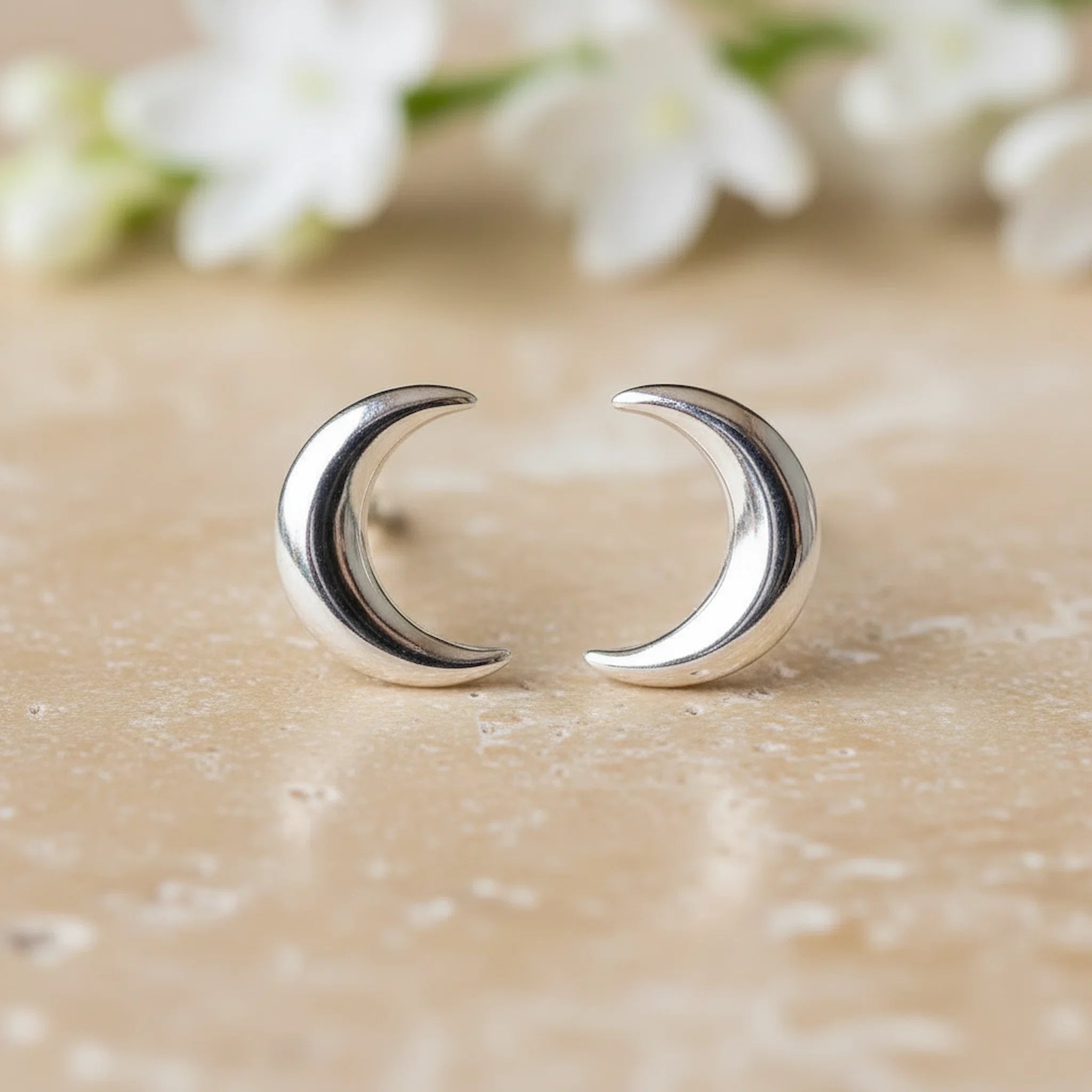 Silver crescent moon earrings on a textured surface with blurred flowers in the background
