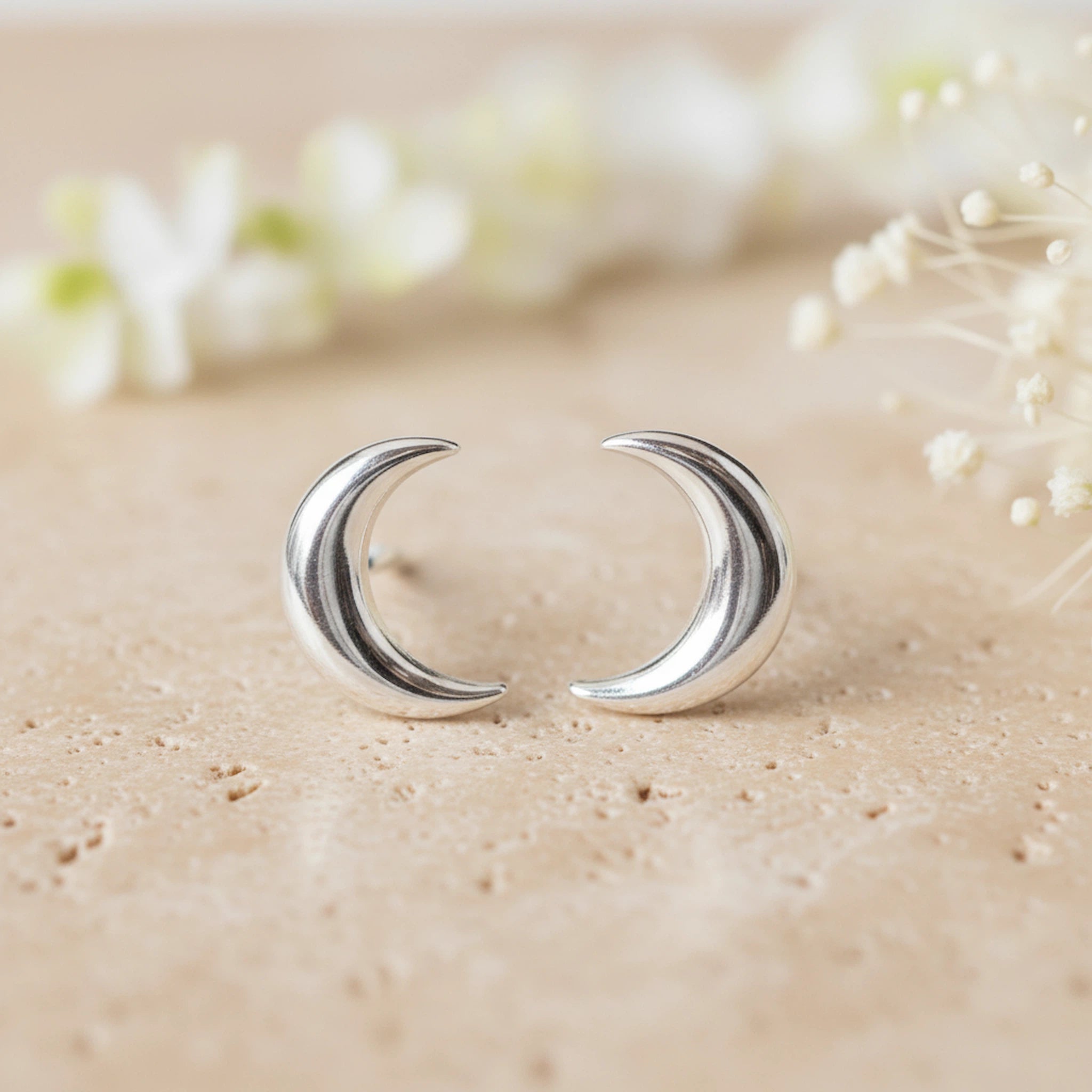 Silver crescent moon earrings on a beige surface with blurred flowers in the background