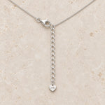 A silver chain necklace with a lobster clasp and an adjustable extension chain ending in a small heart charm engraved with 'i+h', displayed on a light beige stone surface.