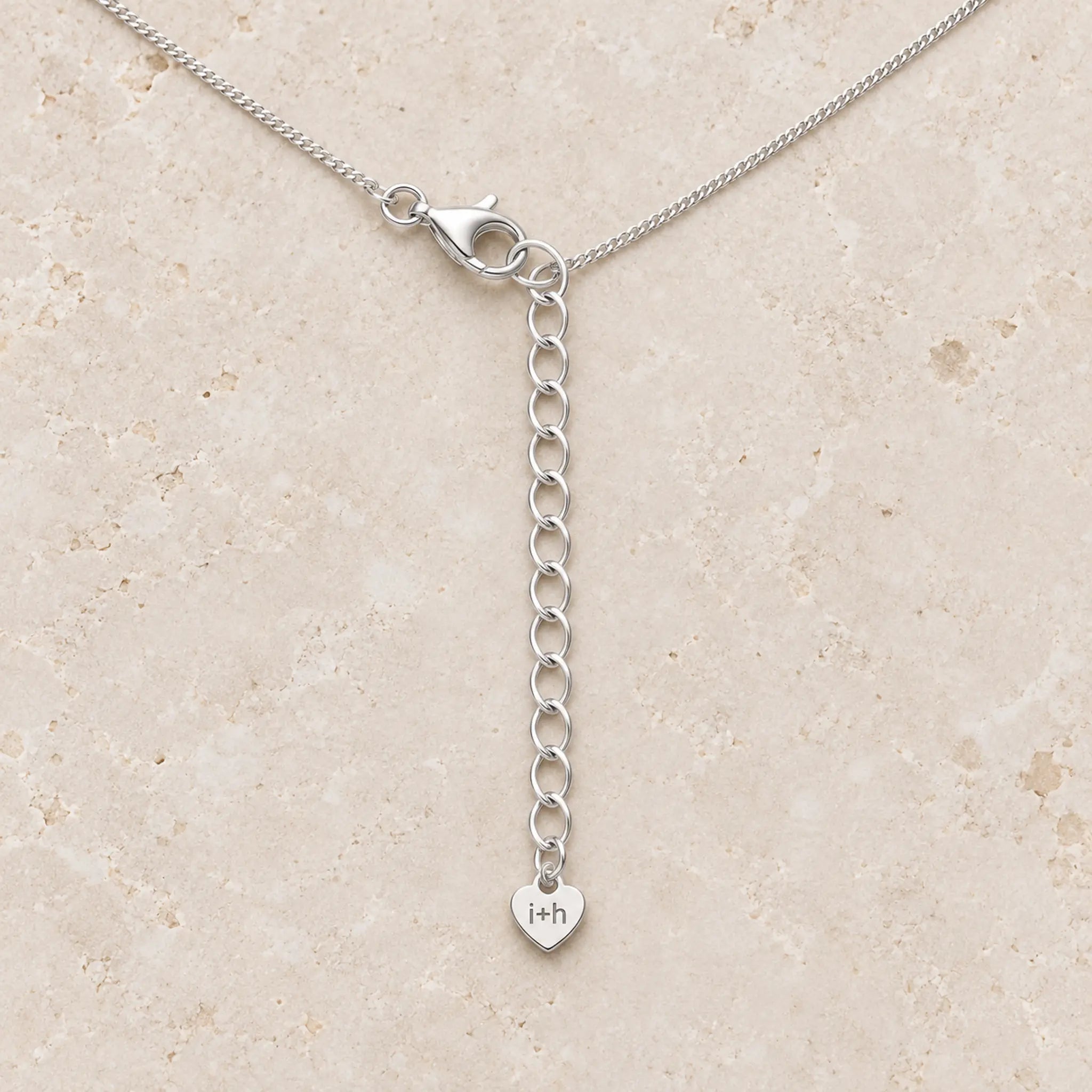 A silver chain necklace with a lobster clasp and an adjustable extension chain ending in a small heart charm engraved with 'i+h', displayed on a light beige stone surface.