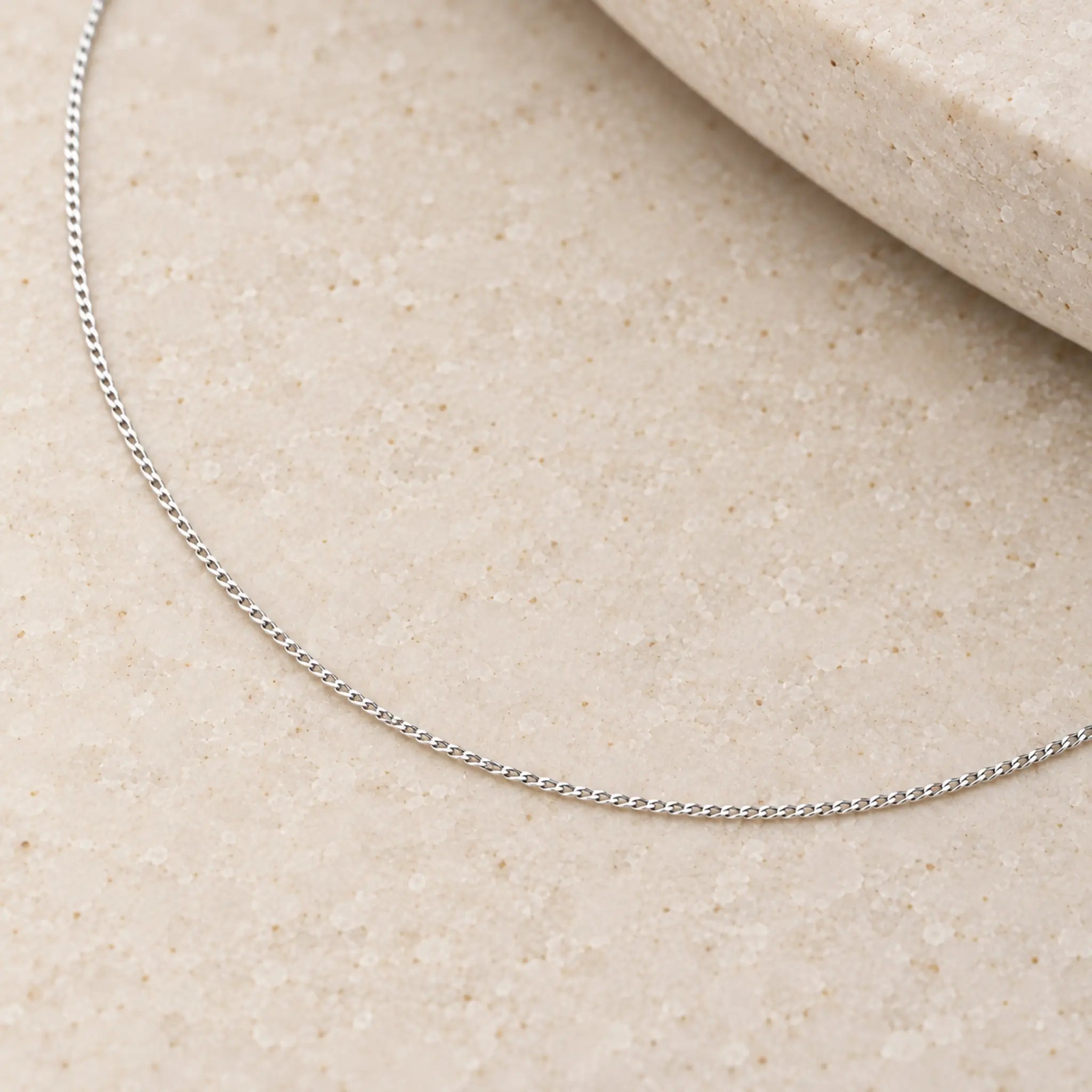 A delicate sterling silver fine 1mm curb chain necklace laid out in a curved line on a light beige textured stone surface.