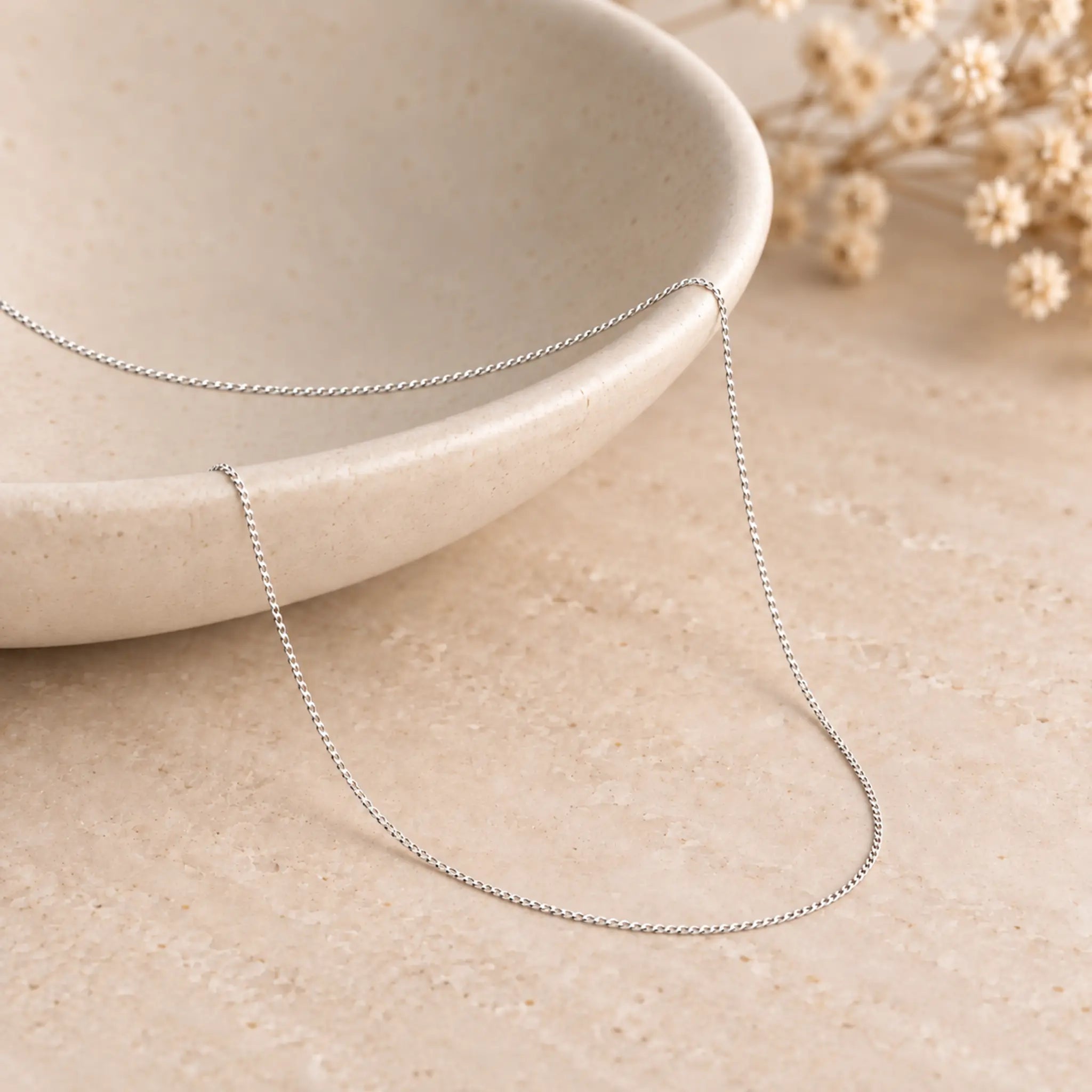 A delicate sterling silver curb chain necklace draped over a light beige ceramic bowl on a textured beige surface, with dried flowers in the background.