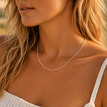 A close-up lifestyle shot of a model wearing a delicate sterling silver fine curb chain necklace, showing the jewelry on her neck against warm natural sunlight.