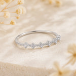 A silver band ring featuring a row of small clear gemstones in a floral or starburst pattern, photographed on a soft beige textured surface with blurred dried flowers in the background.