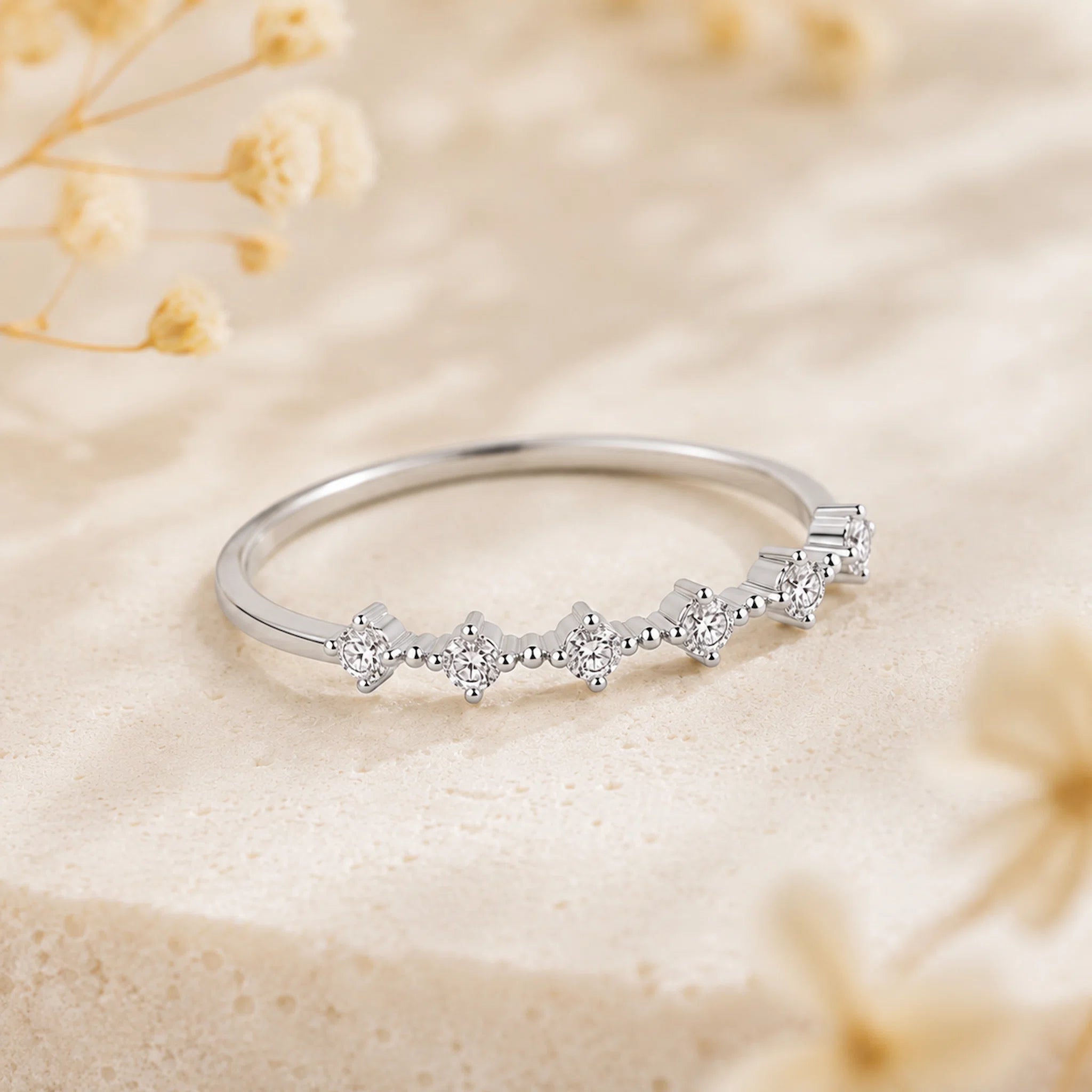 A silver band ring featuring a row of small clear gemstones in a floral or starburst pattern, photographed on a soft beige textured surface with blurred dried flowers in the background.