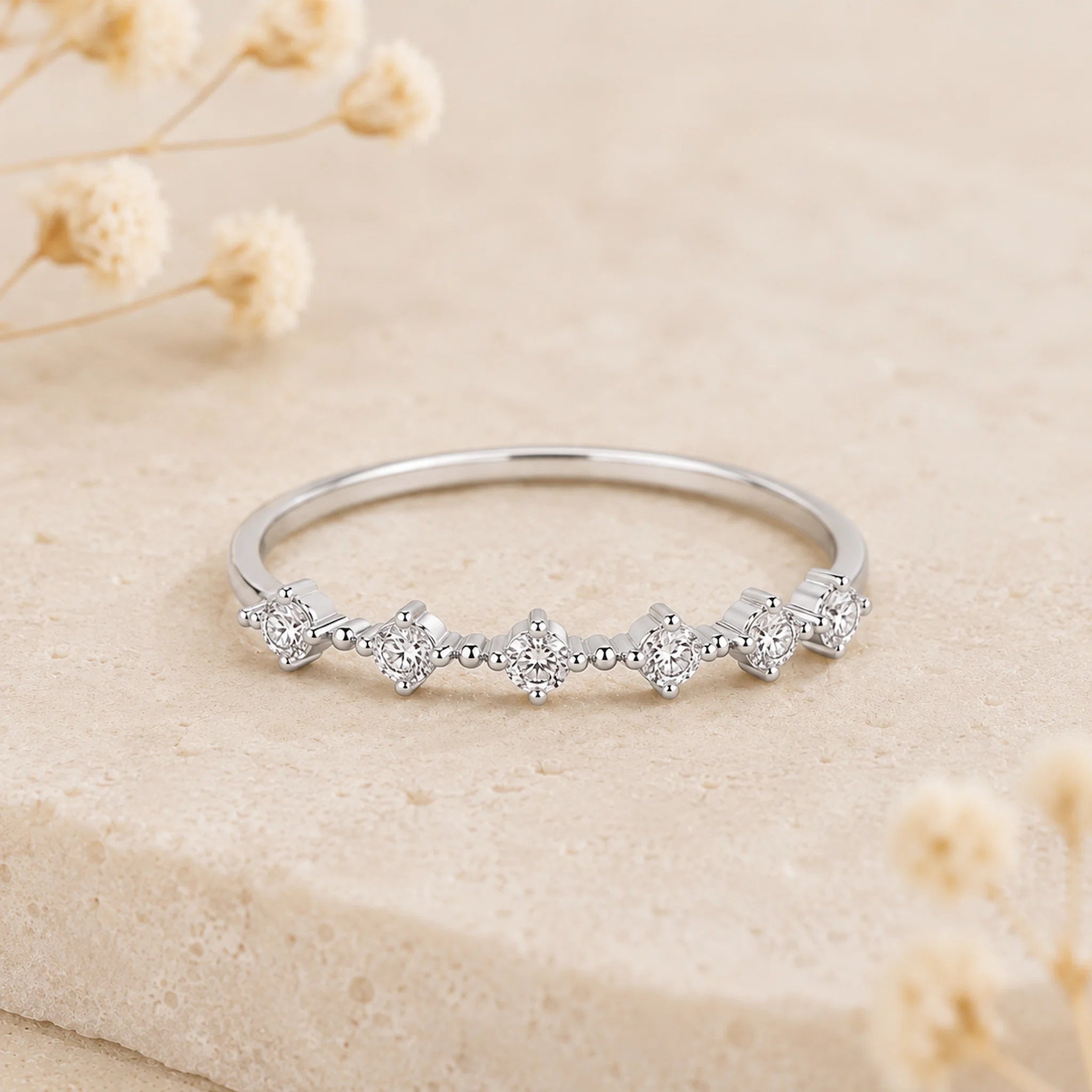 A delicate silver ring featuring five small floral clusters of clear stones, displayed on a textured beige stone surface with dried flowers in the background.