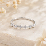 A silver band ring with a row of small clear gemstones in a floral cluster setting, resting on a textured beige stone surface with soft dried flowers in the background.