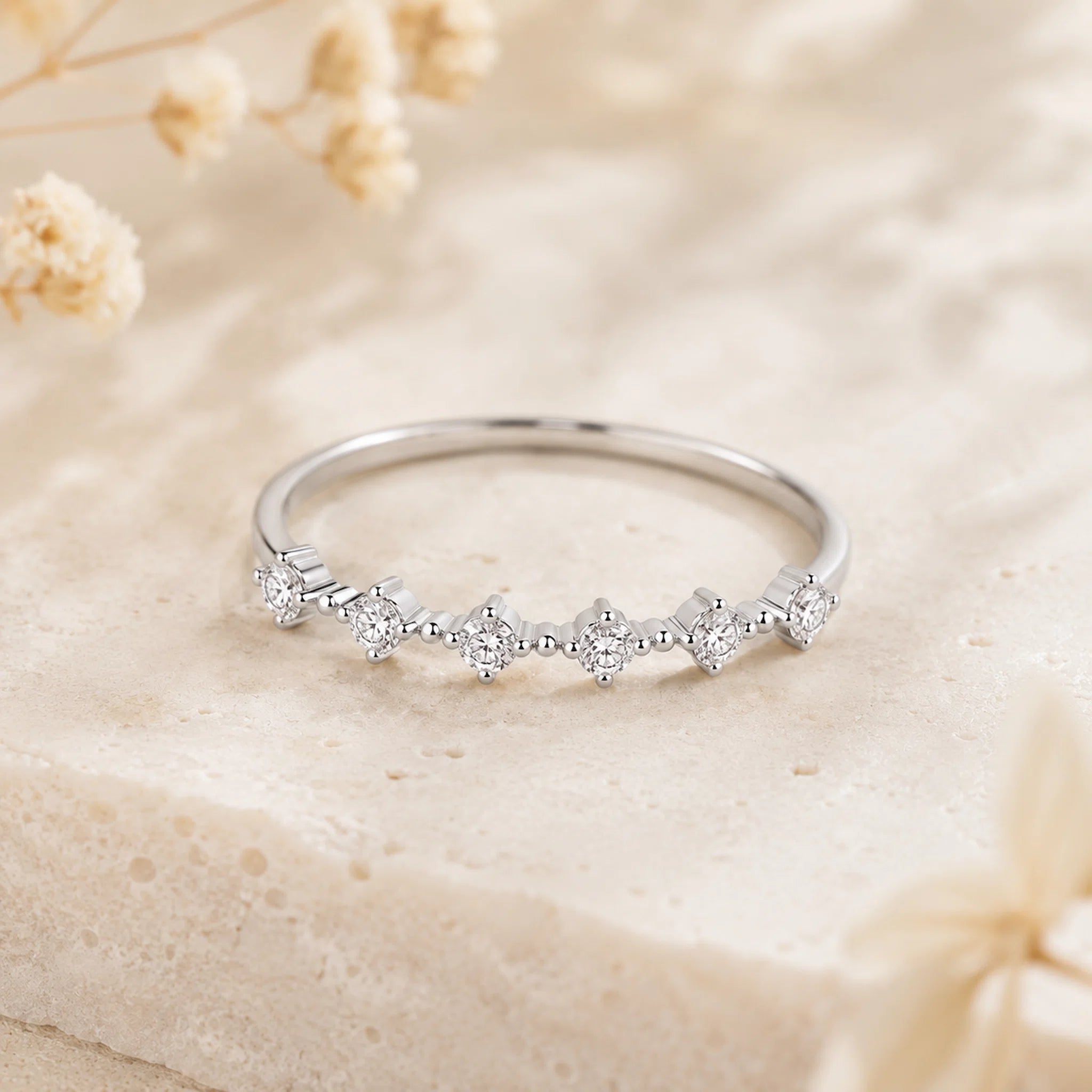 A silver band ring with a row of small clear gemstones in a floral cluster setting, resting on a textured beige stone surface with soft dried flowers in the background.