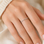 A delicate silver ring with small clear stones worn on a person's ring finger, shown in a close-up lifestyle shot against a soft cream fabric background.