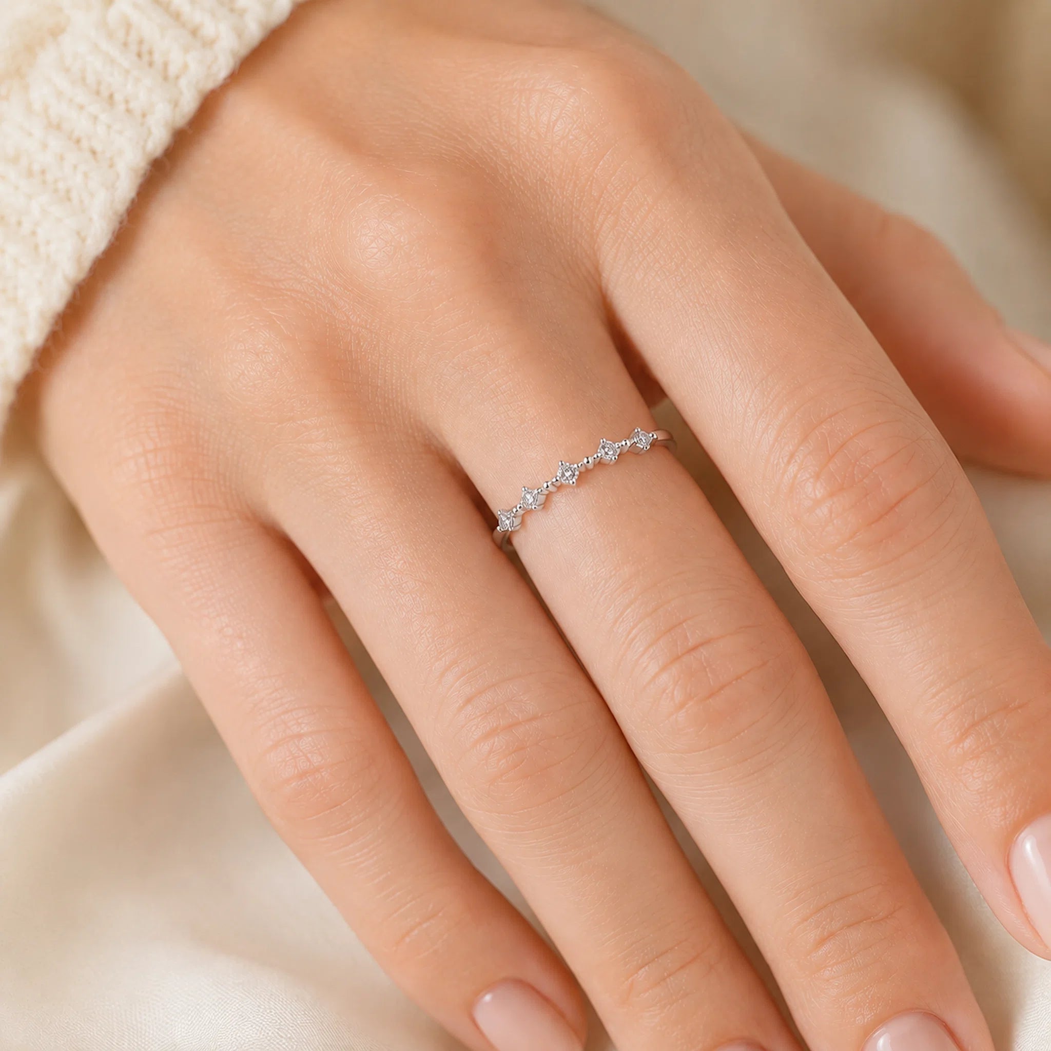A delicate silver ring with small clear stones worn on a person's ring finger, shown in a close-up lifestyle shot against a soft cream fabric background.