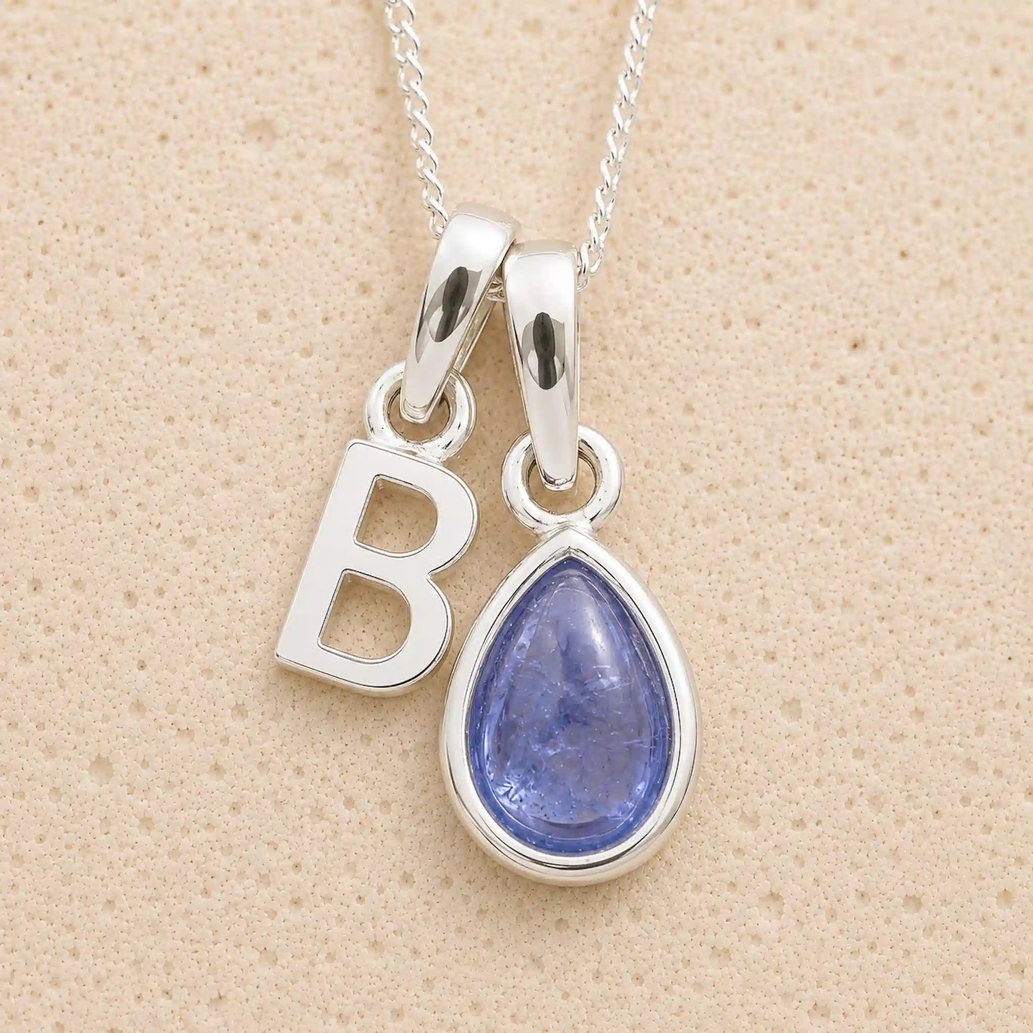 Silver december birthstone necklace with 'B' pendant and teardrop-shaped tanzanite gemstone on beige background