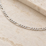 A silver Figaro chain necklace displayed on a textured beige stone surface.