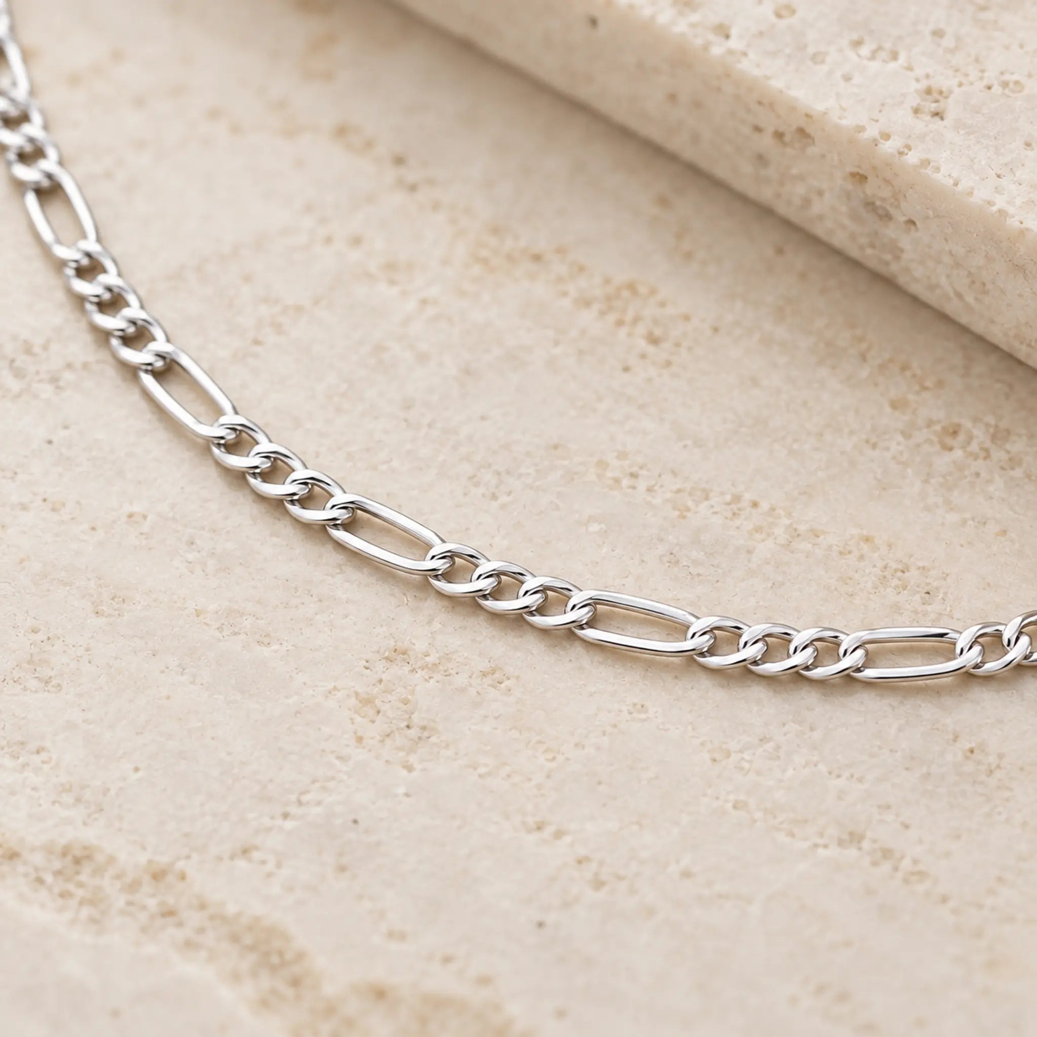 A silver Figaro chain necklace displayed on a textured beige stone surface.