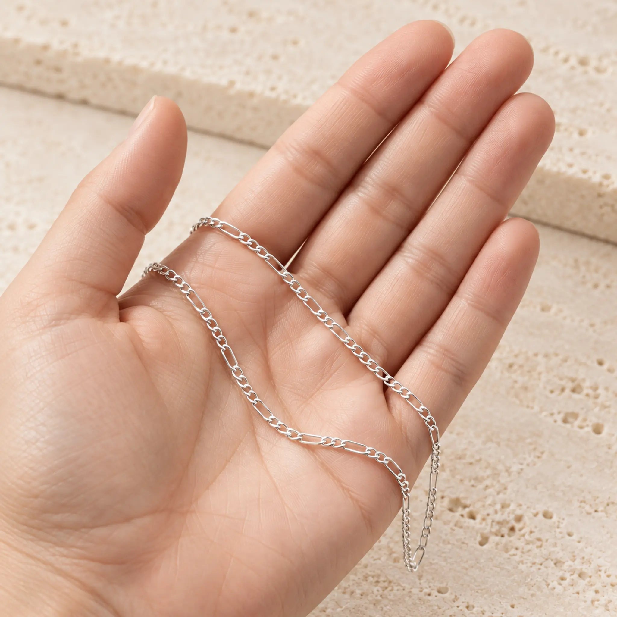 A delicate silver chain necklace draped across an open hand, shown against a light beige textured stone background.