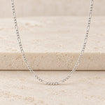 A silver Figaro chain necklace displayed on a textured beige stone surface.