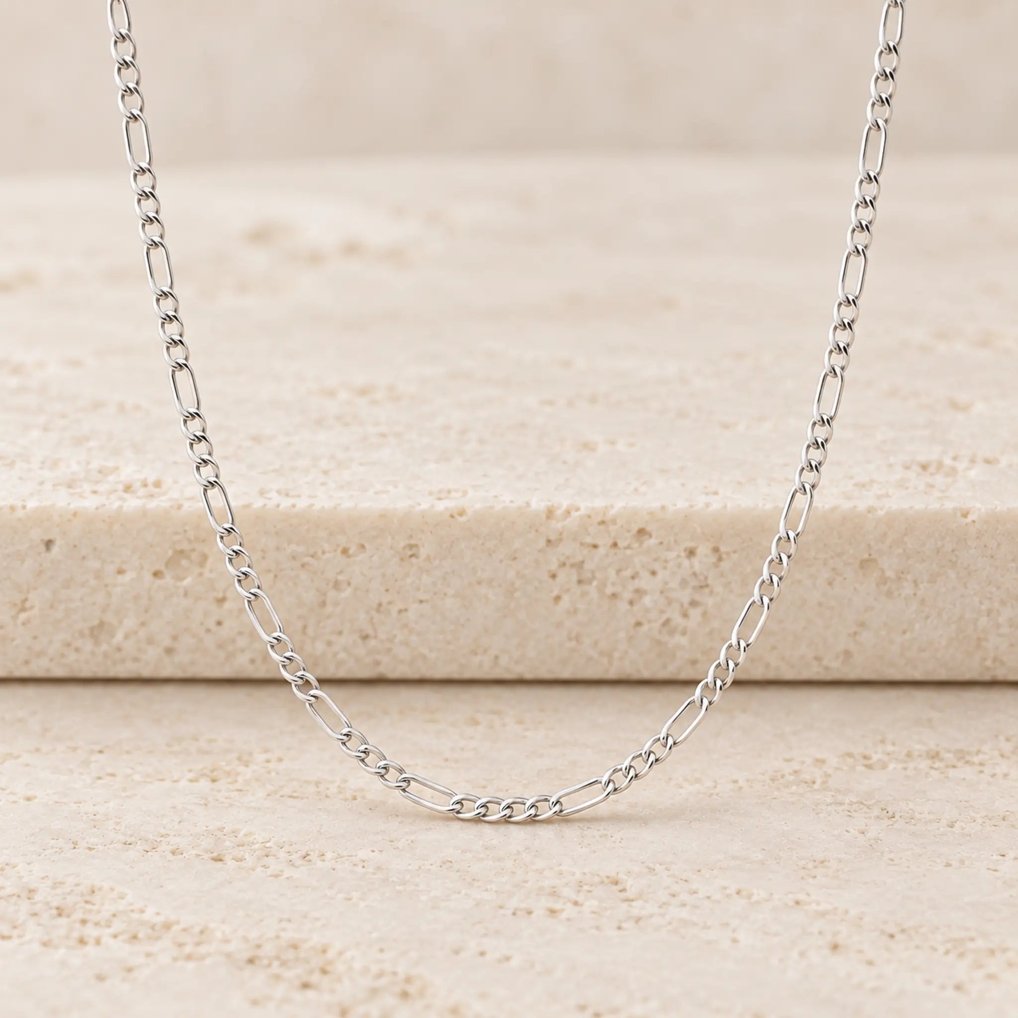 A silver Figaro chain necklace displayed on a textured beige stone surface.