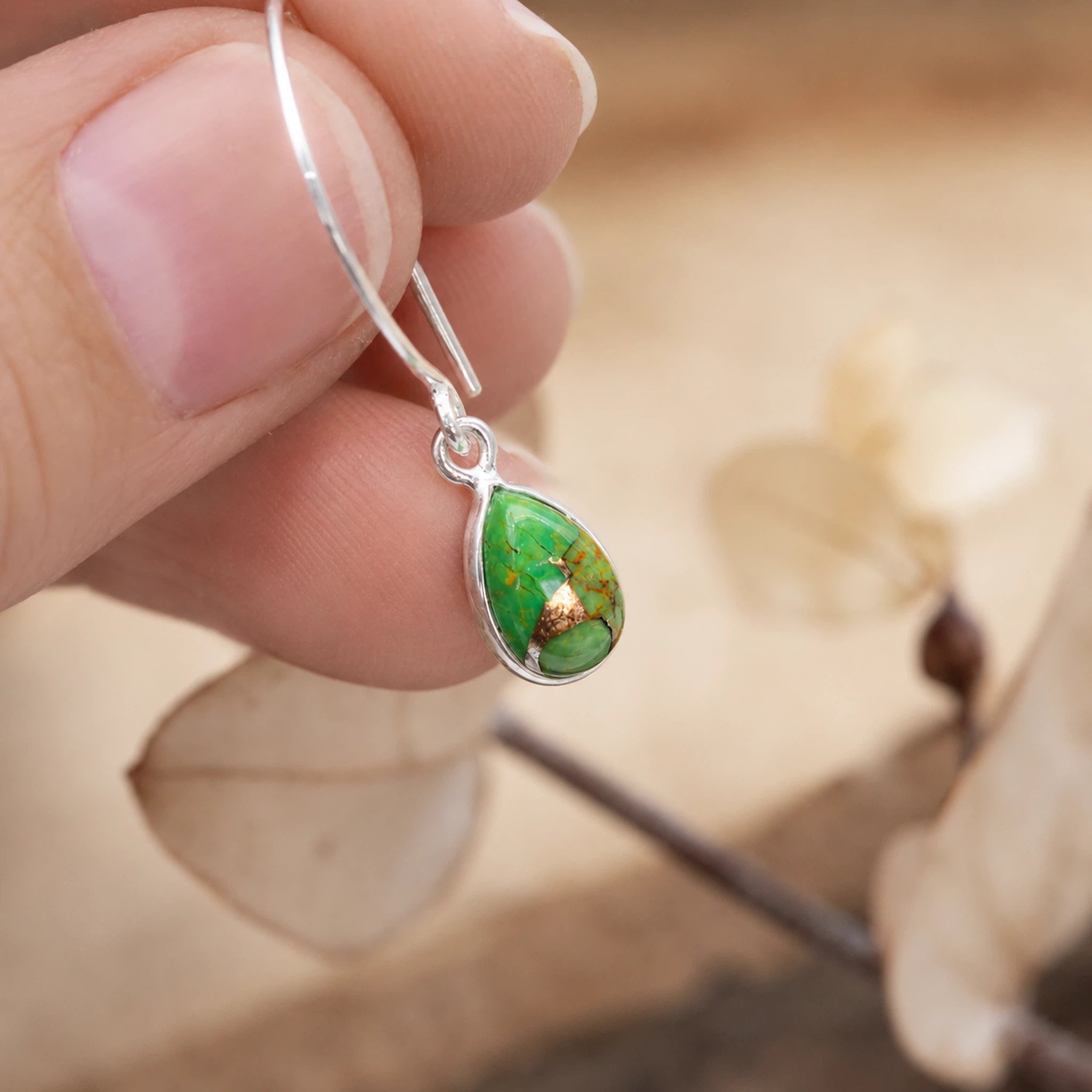 Green teardrop earring held between fingers with a blurred natural background
