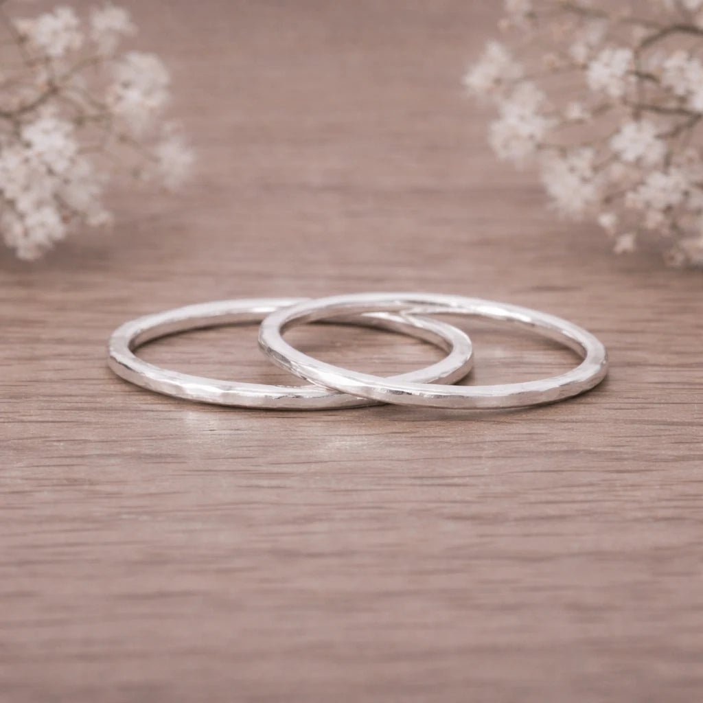 Two silver hammered stacker rings on a wooden surface with blurred flowers in the background