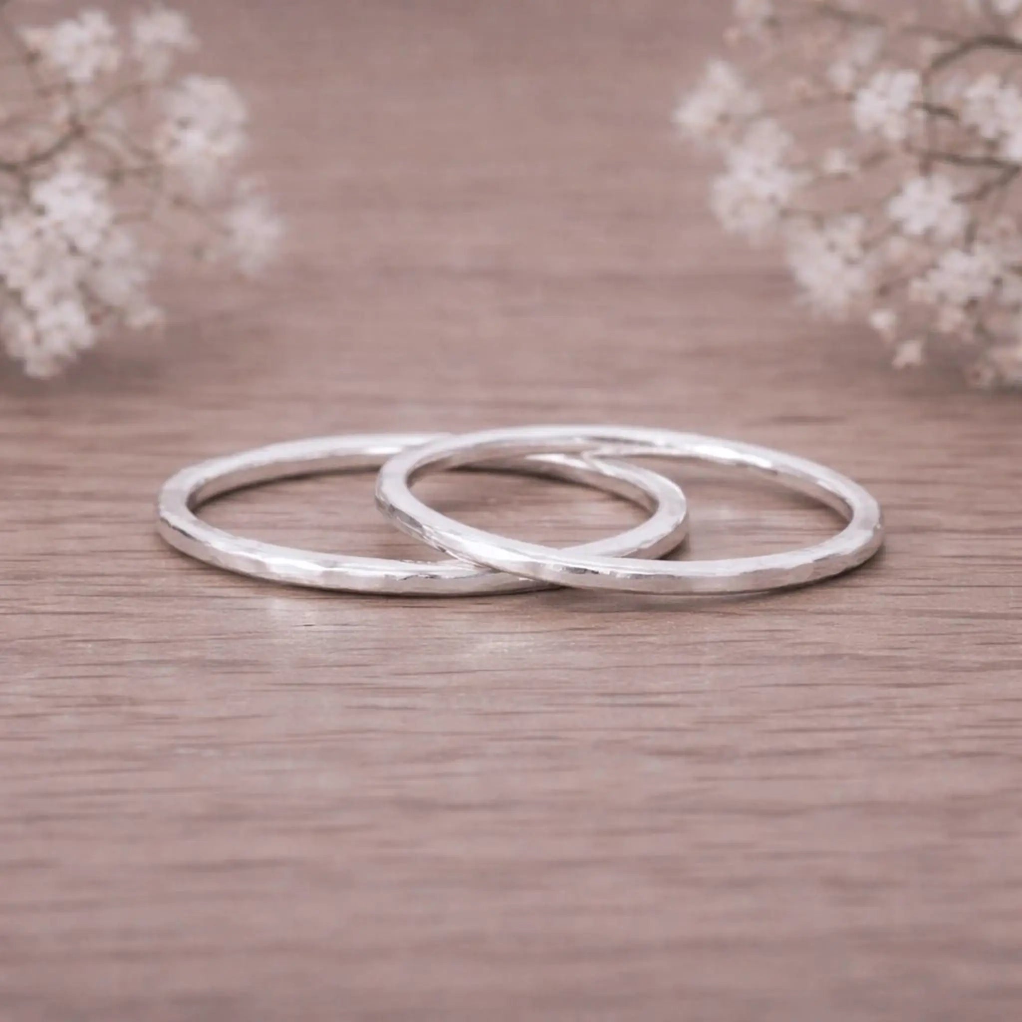 Two sterling silver hammered stacker rings on a wooden surface with blurred flowers in the background