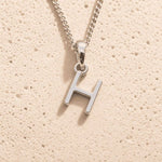 Sterling Silver Initial Necklace with a minimalist H charm pendant on a light stone background