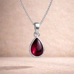 Teardrop-shaped red natural garnet gemstone pendant on a sterling silver chain against a beige background