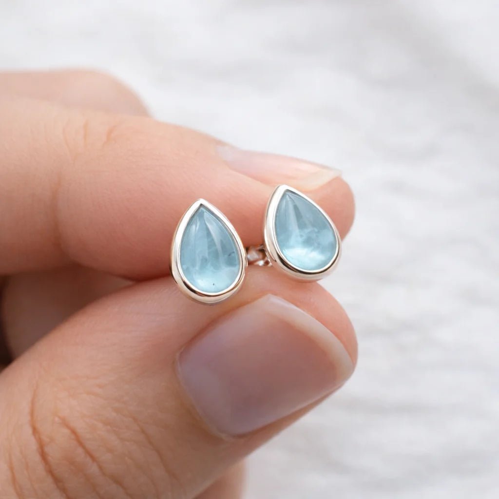 Teardrop-shaped earrings with light blue natural aquamarine gemstones held between fingers against a blurred background