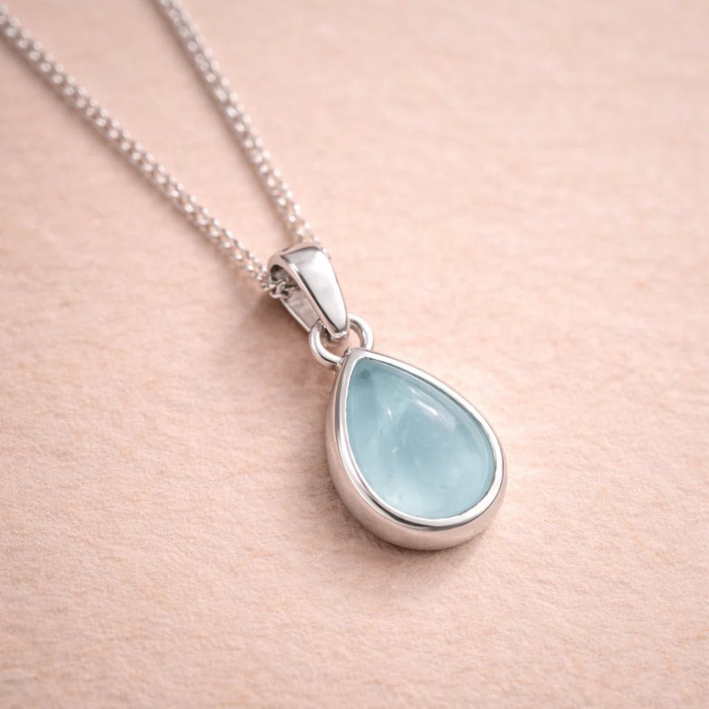 Teardrop-shaped pendant with light blue natural aquamarine stone on a sterling silver chain against a beige background