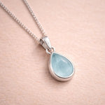 Teardrop-shaped pendant with light blue natural aquamarine stone on a sterling silver chain against a beige background