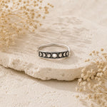 A silver moon phase ring with black and white inlays, displayed on a textured beige stone surrounded by dried flowers.