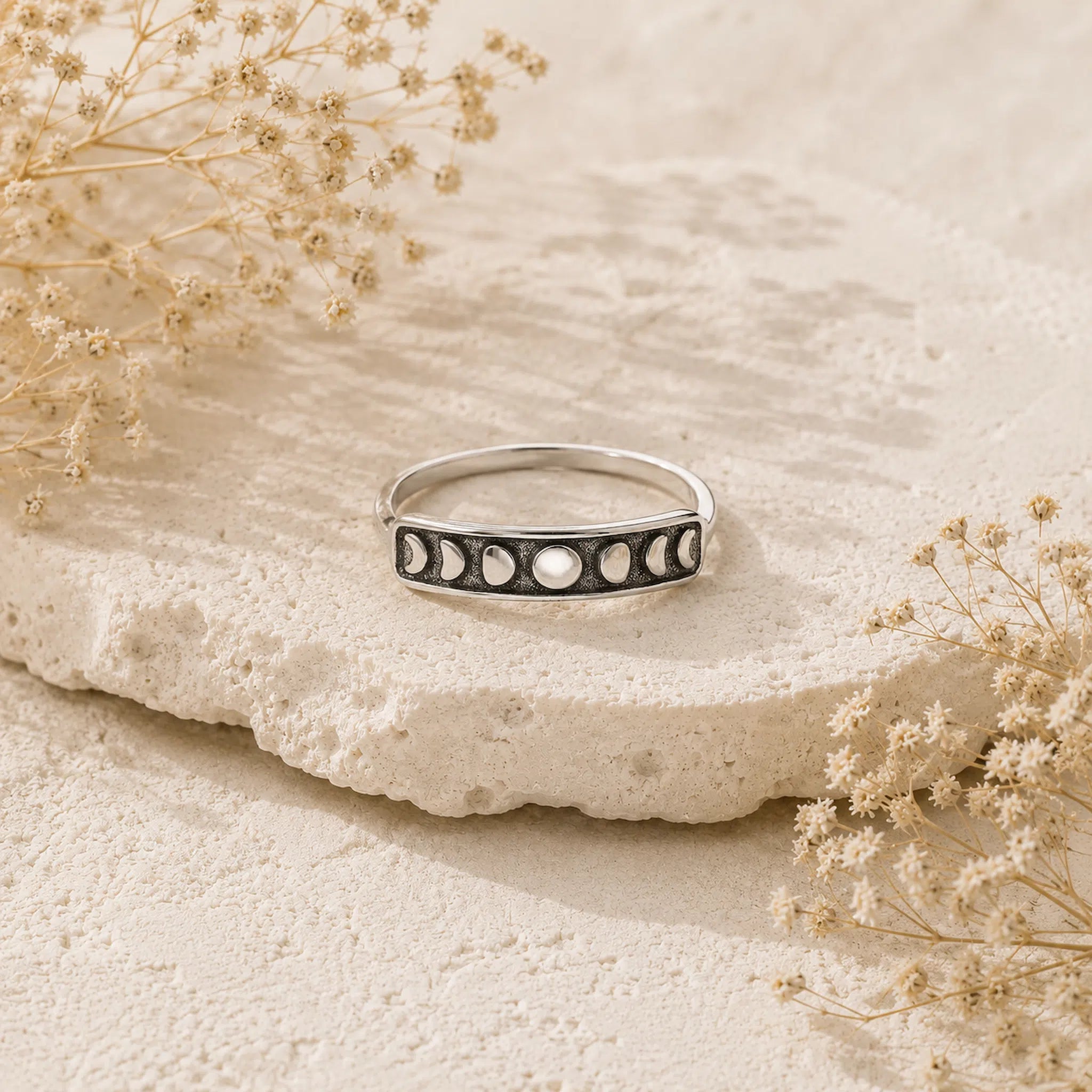 A silver moon phase ring with black and white inlays, displayed on a textured beige stone surrounded by dried flowers.