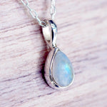 A silver teardrop pendant moonstone necklace with an iridescent moonstone gemstone, resting on a light-colored wooden surface.