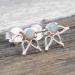 A pair of silver stud earrings with natural australian opals, resting on a rustic wooden surface. The opal stones exhibit iridescent colours, and the silver posts have a delicate, lotus openwork design.
