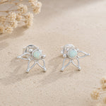 A pair of silver stud earrings with round natural australia opal center, displayed on a textured beige surface with dried flowers.