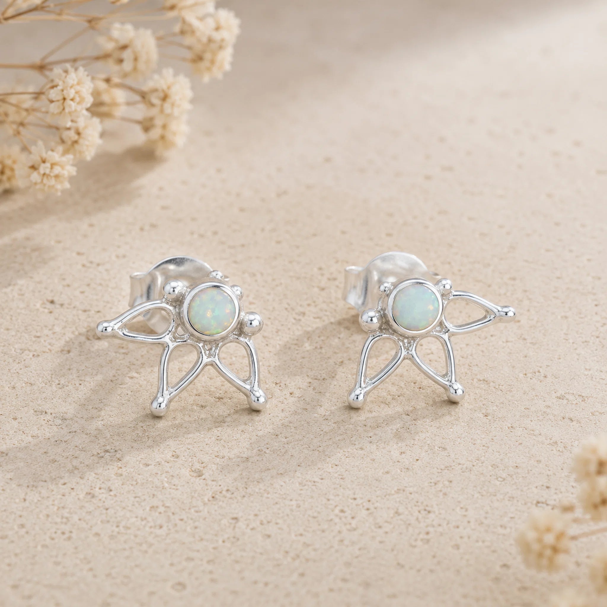 A pair of silver stud earrings with round natural australia opal center, displayed on a textured beige surface with dried flowers.